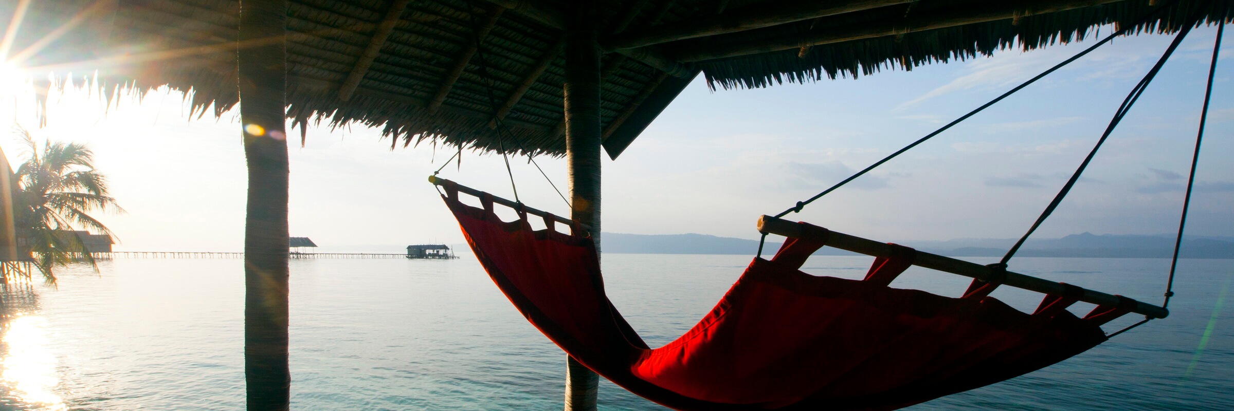 Zwei Gäste entspannen in einer roten Hängematte auf der Terrasse eines Overwater-Bungalows im Kri Eco Resort. Sie genießen den direkten Blick auf das ruhige türkisfarbene Meer und die untergehende Sonne, während ein weiterer Gast im Wasser schwimmt.