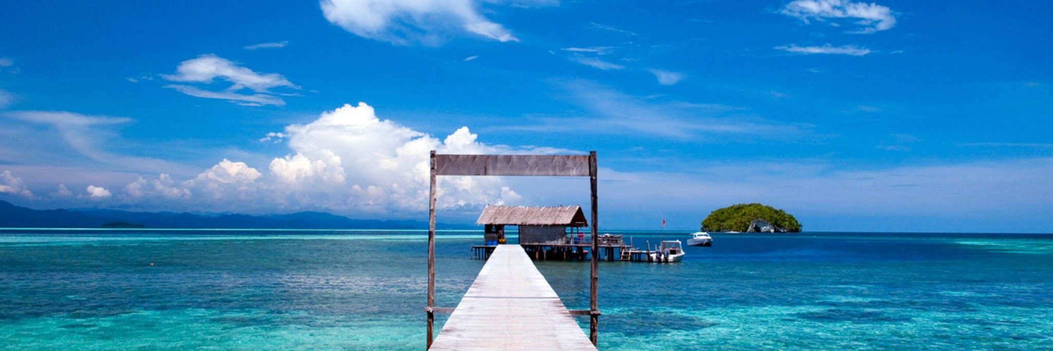 Ein langer, hölzerner Bootssteg des Sorido Bay Resorts führt vom Privatstrand weit über das klare türkisfarbene Wasser der Lagune. Im Hintergrund sind weitere Bungalows, Palmen und die tropische Vegetation der Insel Kri zu sehen.