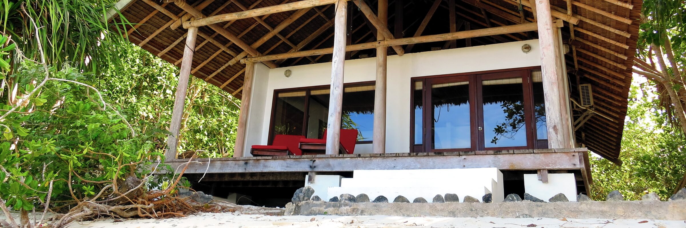 Eine Reihe von drei traditionellen Strand-Bungalows des Sorido Bay Resorts, auf Stelzen direkt im weißen Sand unter Kokospalmen. Der Weg davor ist mit einem Steg gesäumt, das klare türkisfarbene Meer liegt direkt davor.