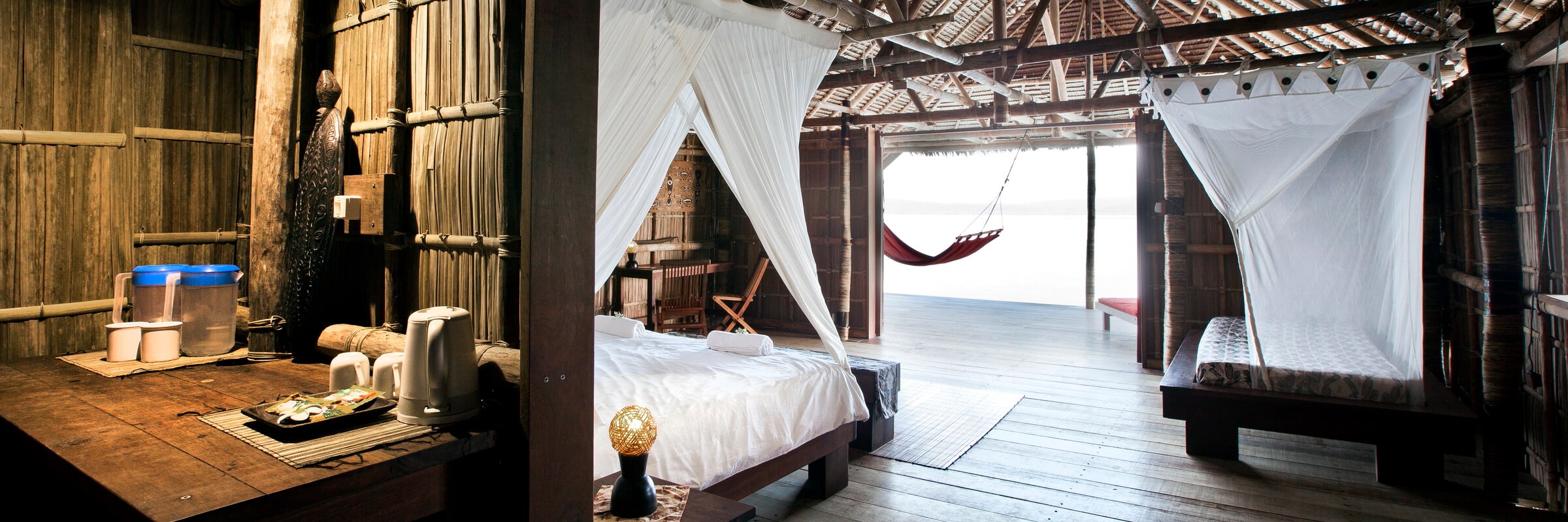 Innenansicht eines komfortablen Schlafzimmers in einem Overwater-Bungalow des Kri Eco Resort. Es zeigt ein bequemes Doppelbett mit Moskitonetz, Holzverkleidung an den Wänden, eine Holzterrasse mit einer Hängematte und einen atemberaubenden Blick auf das türkisfarbene Meer.