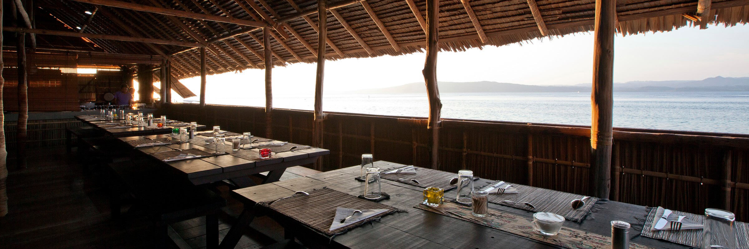 Innenansicht des halboffenen Restaurants des Kri Eco Resort bei Sonnenuntergang. Lange Holztische und Bänke sind für das Abendessen gedeckt. Der Blick geht auf das ruhige Meer und den Horizont, mit Bergen in der Ferne.