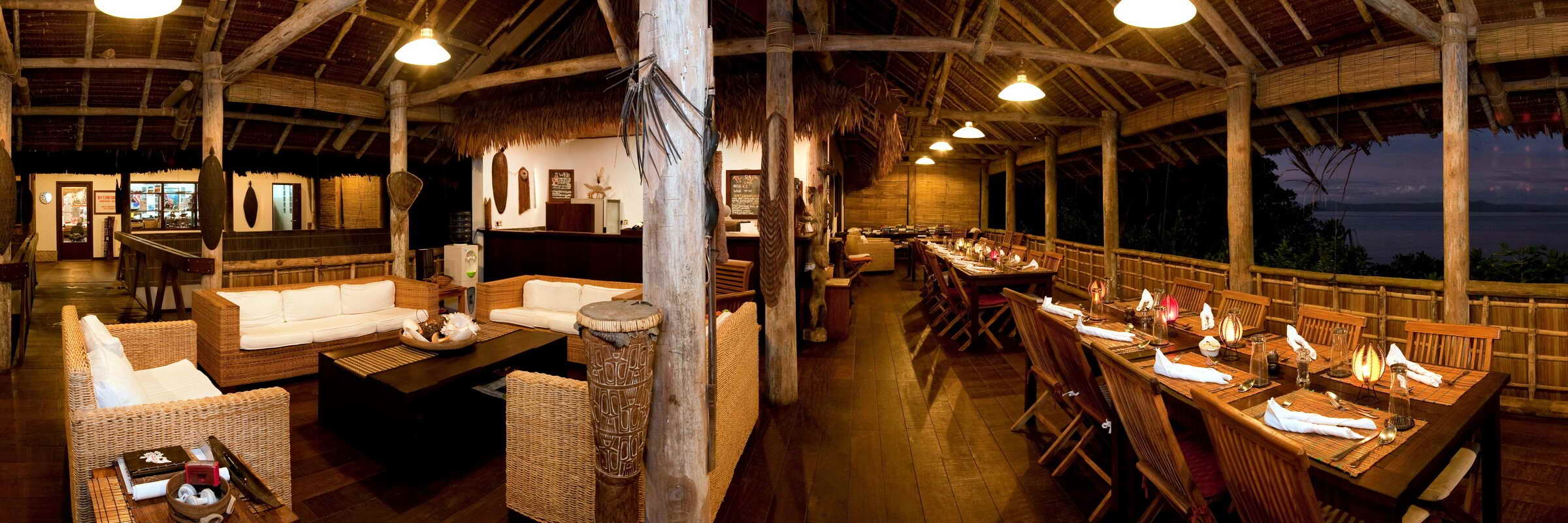 Innenansicht des gemütlichen, halboffenen Restaurants im Sorido Bay Resort. Mehrere Holz-Esstische sind unter einem hohen, traditionell gedeckten Dach angeordnet, mit einem herrlichen Blick auf die Lagune und das offene Meer bei Tageslicht.