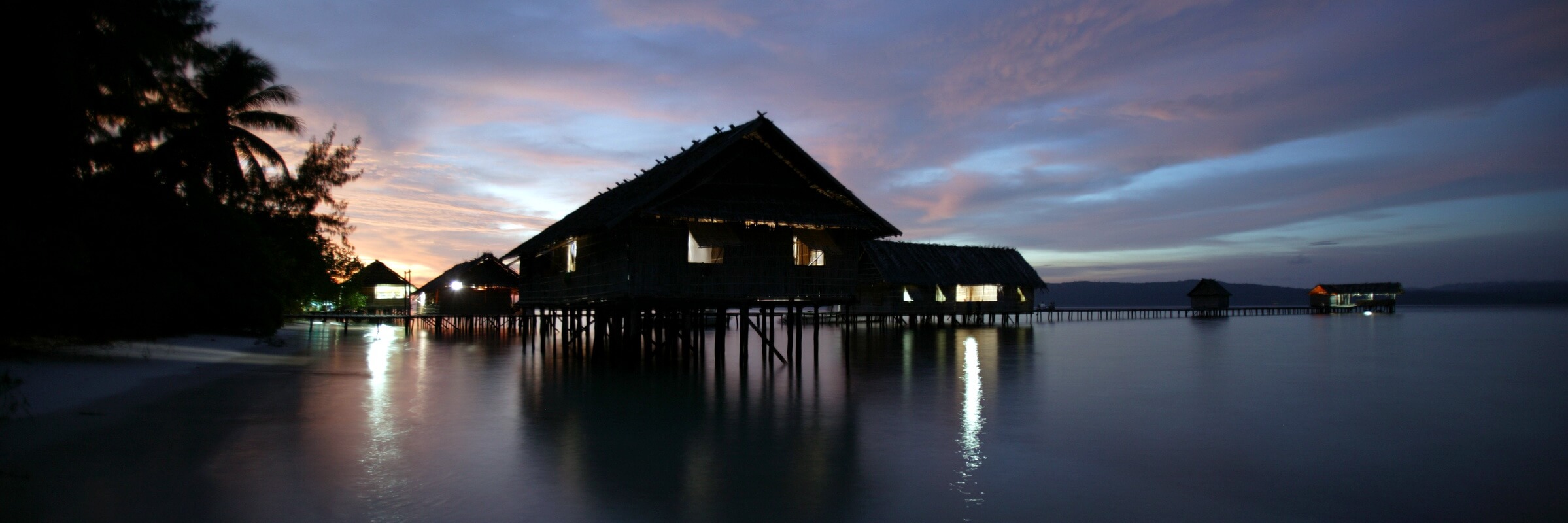 Nachtaufnahme einer Reihe beleuchteter, traditioneller Overwater-Bungalows des Kri Eco Resort auf Stelzen über dem dunklen Wasser von Raja Ampat. Ein langer Holzsteg führt zu den Bungalows. Die Beleuchtung spiegelt sich im Wasser.