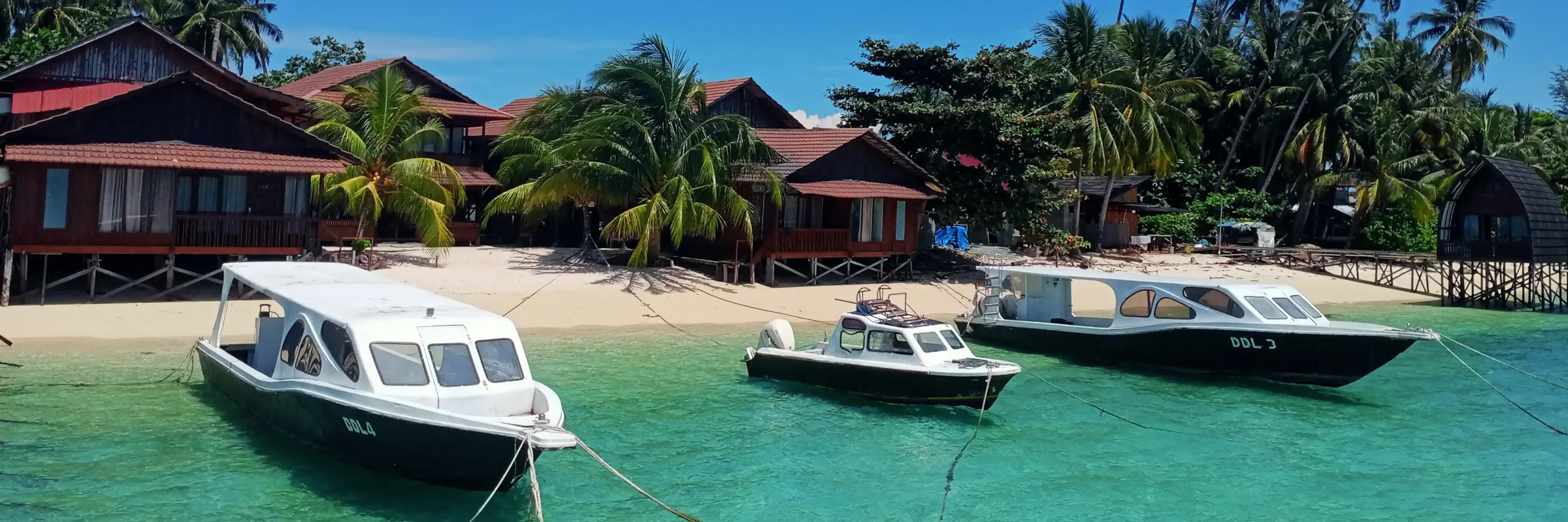 Tauchboote der Derawan Dive Lodge am weißen Sandstrand von Pulau Derawan in Ost-Kalimantan, Indonesien, vor türkisfarbenem Meer.