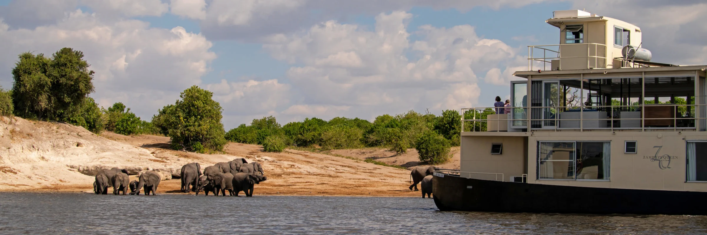 Das luxuriöse Hausboot Zambezi Queen ankert am Ufer, während eine Herde Elefanten am Fluss trinkt.