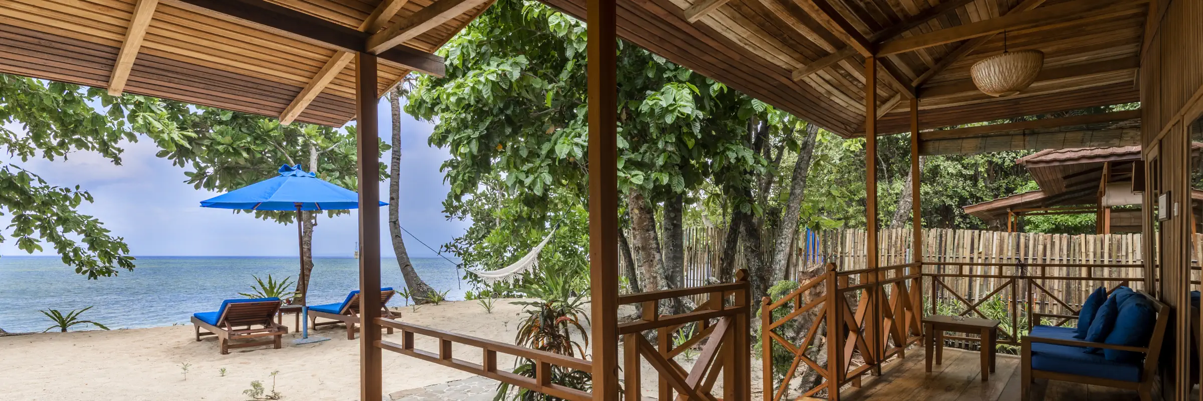 Blick von der schattigen Holzterrasse einer Beach View Villa auf den weißen Sandstrand mit blauem Sonnenschirm und das ruhige Meer auf Siladen.