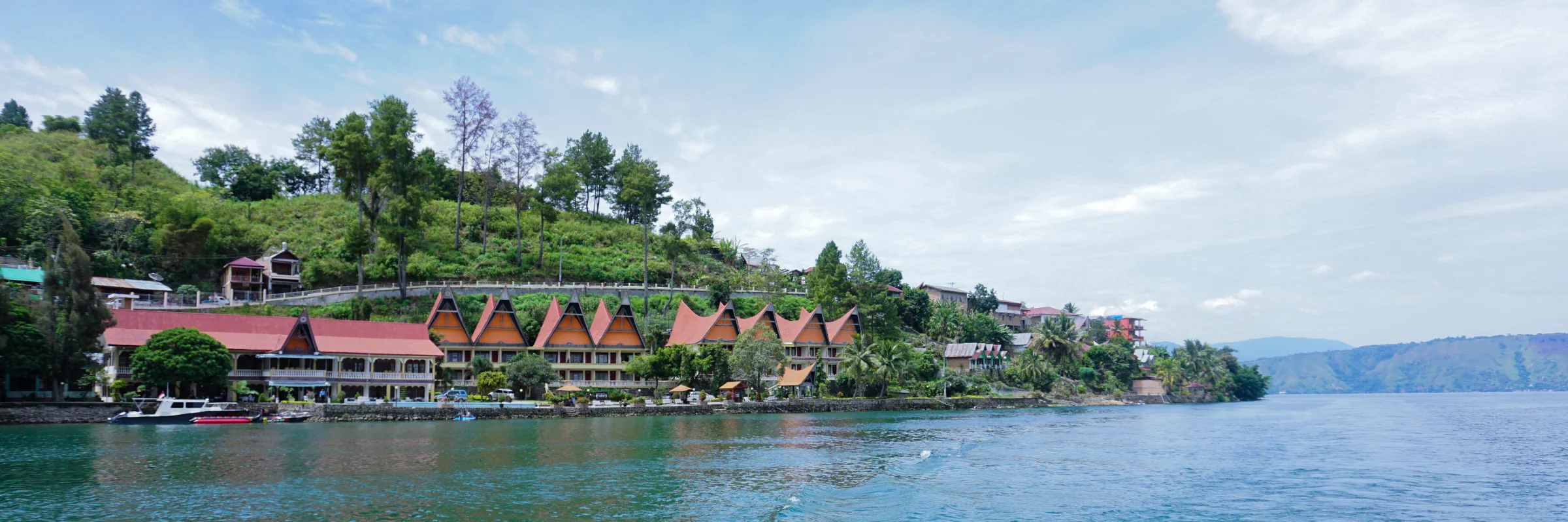 Blick vom Wasser auf die Küste der Insel Samosir am Toba-See auf Sumatra mit traditionellen Batak-Häusern und Hotels vor grünen Hügeln.