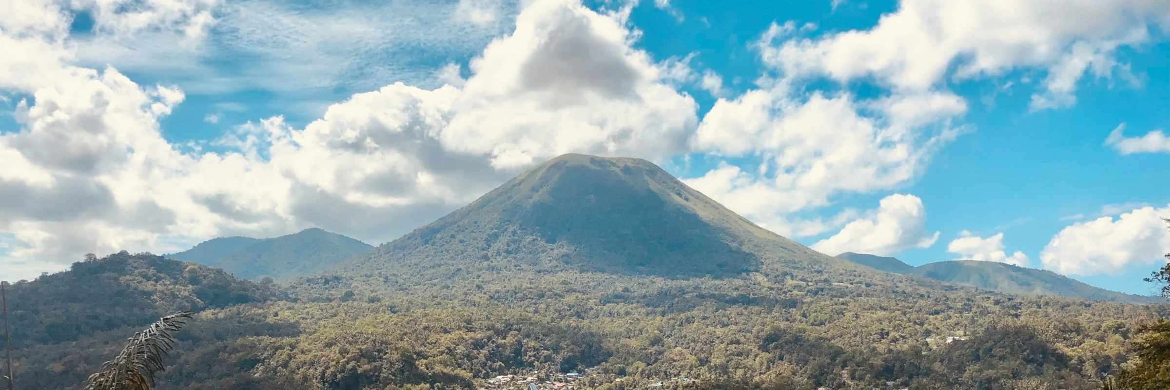 Der bewaldete Vulkan Lokon in der Region Minahasa auf Sulawesi, umgeben von tropischer Berglandschaft unter blauem Himmel mit weißen Wolken.