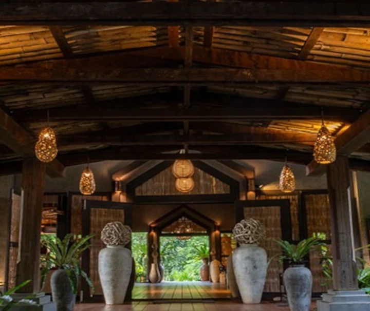 Offene, traditionelle Holz-Lobby des Nam Kat Yorla Pa Luxus-Öko-Resorts in Laos mit geflochtenen Hängeleuchten und tropischen Pflanzen.