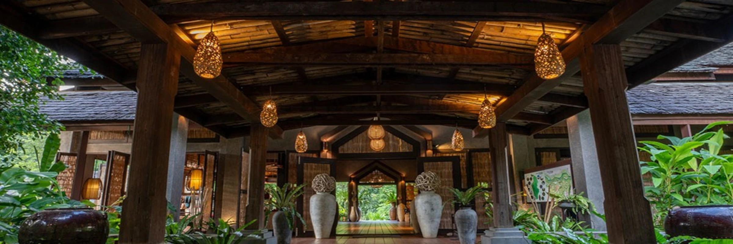 Offene, traditionelle Holz-Lobby des Nam Kat Yorla Pa Luxus-Öko-Resorts in Laos mit geflochtenen Hängeleuchten und tropischen Pflanzen.