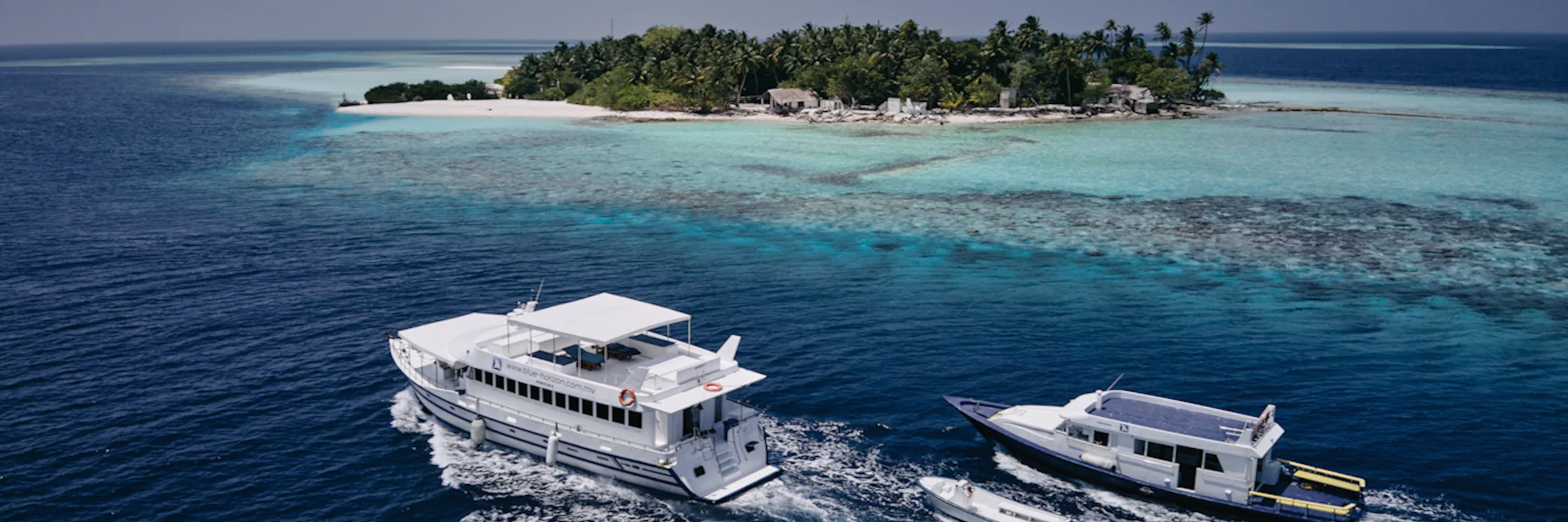 Das Safari-Boot Horizon II navigiert an einem flachen Korallenriff und einer palmengesäumten Tropeninsel auf den Malediven vorbei.