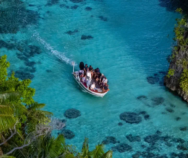 Ein Expeditions-Beiboot mit Urlaubern fährt durch glasklares, türkisfarbenes Wasser vorbei an tropischen Palmen und Felsen in Raja Ampat.