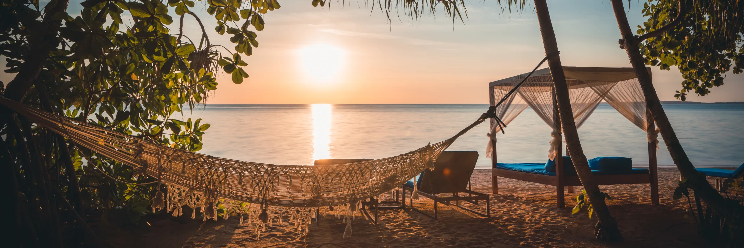Eine weiße Hängematte und ein Strandbett aus Holz am Sandstrand von Siladen bei einem idyllischen Sonnenuntergang über dem Meer.