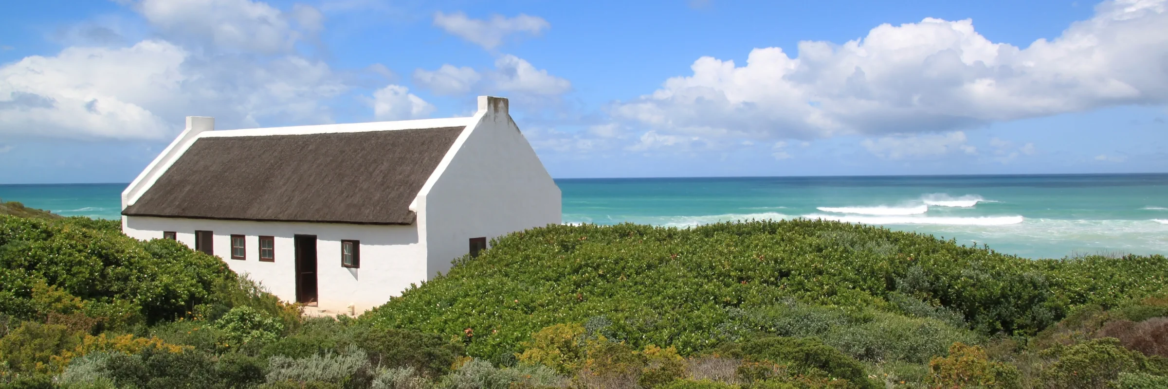 Reetgedecktes weißes Fischerhaus in den Dünen vor dem hellblauen Meer in Arniston