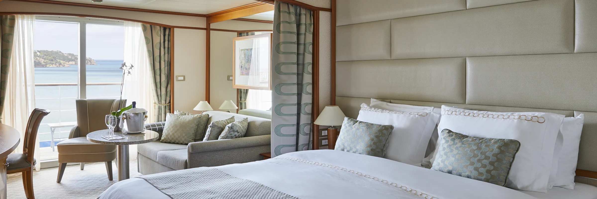 Innenaufnahme einer geräumigen und luxuriösen Kreuzfahrtschiff-Suite mit einem großen Bett, einem Sitzbereich, einem Esstisch und einem Balkon mit Meerblick.