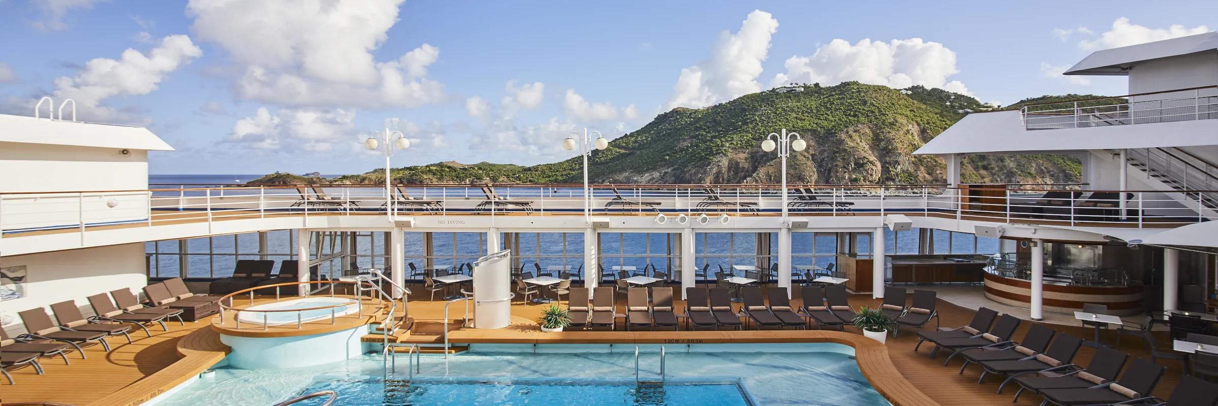 Das Oberdeck der Silversea mit einem Swimmingpool, Whirlpools, Liegestühlen und einem Blick auf eine bergige Inselküste.