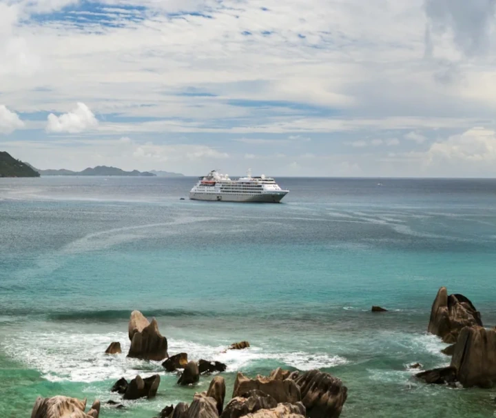 Ein Panorama-Foto einer tropischen Küste mit Granitfelsen im Wasser, türkisfarbenem Meer, einer fernen Insel und einem Kreuzfahrtschiff.