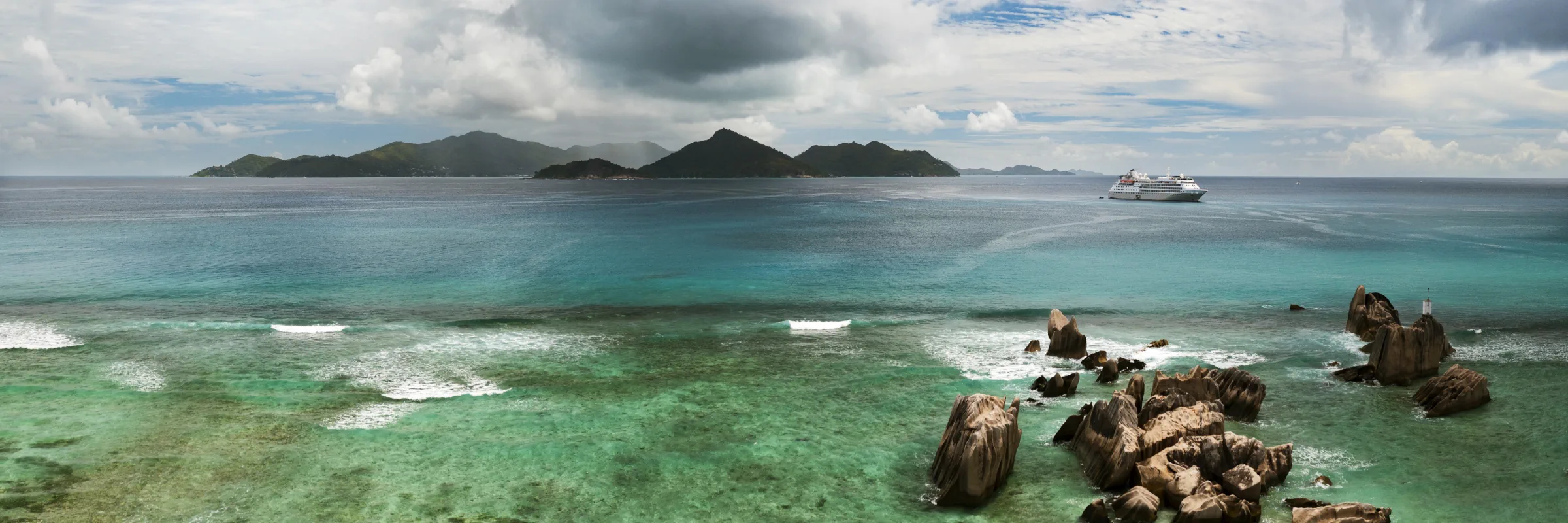 Ein Panorama-Foto einer tropischen Küste mit Granitfelsen im Wasser, türkisfarbenem Meer, einer fernen Insel und einem Kreuzfahrtschiff.
