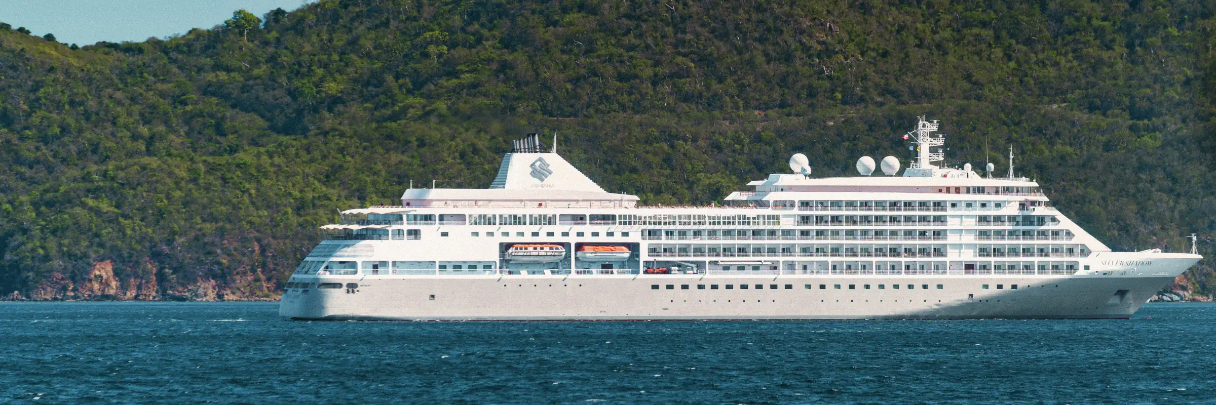 Kreuzfahrtschiff der Silversea-Flotte auf dem blauen Meer vor einer grünen, bergigen Inselküste.