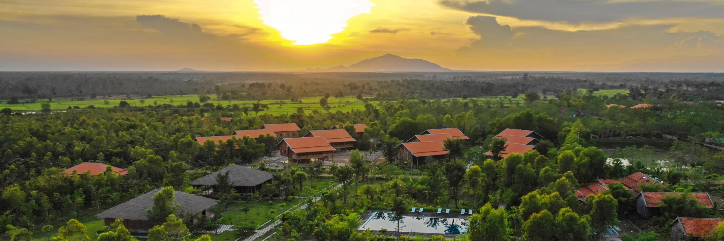 Luftaufnahme des Farmhouse Resort & Spa in Kambodscha bei Sonnenuntergang mit Blick auf grüne Reisfelder und Berge.