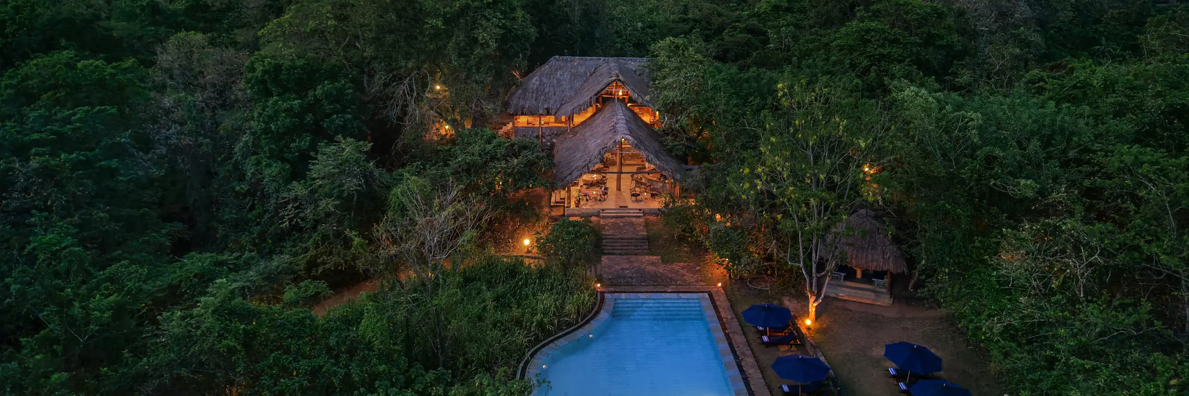 Abendliche Luftaufnahme der Gal Oya Lodge Sri Lanka mit beleuchteten strohgedeckten Naturgebäuden, umgeben von dichtem Dschungel, sowie einem beleuchteten Swimmingpool im Vordergrund.