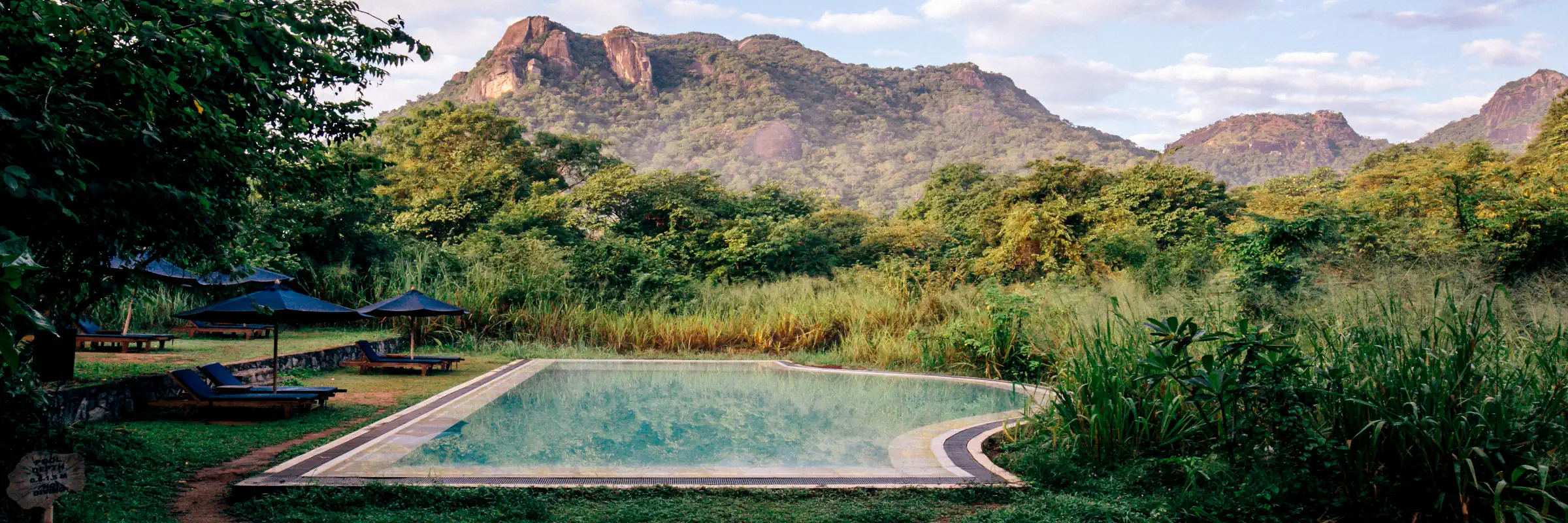Großer Swimmingpool der Gal Oya Lodge Sri Lanka mit Liegen und Sonnenschirmen vor einer üppigen Vegetation und der markanten Bergkulisse des Gal Oya Nationalparks.