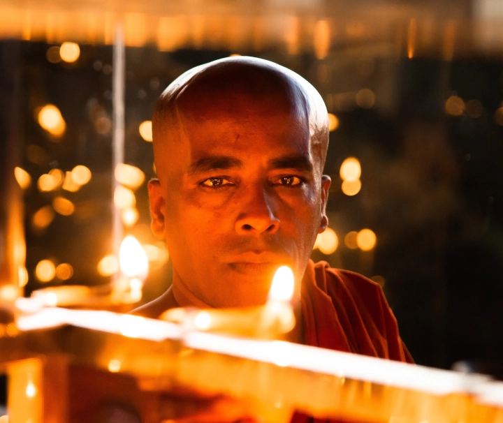 Ein buddhistischer Mönch blickt während einer traditionellen Pooja-Zeremonie in Sri Lanka hinter leuchtenden Öllampen hervor.