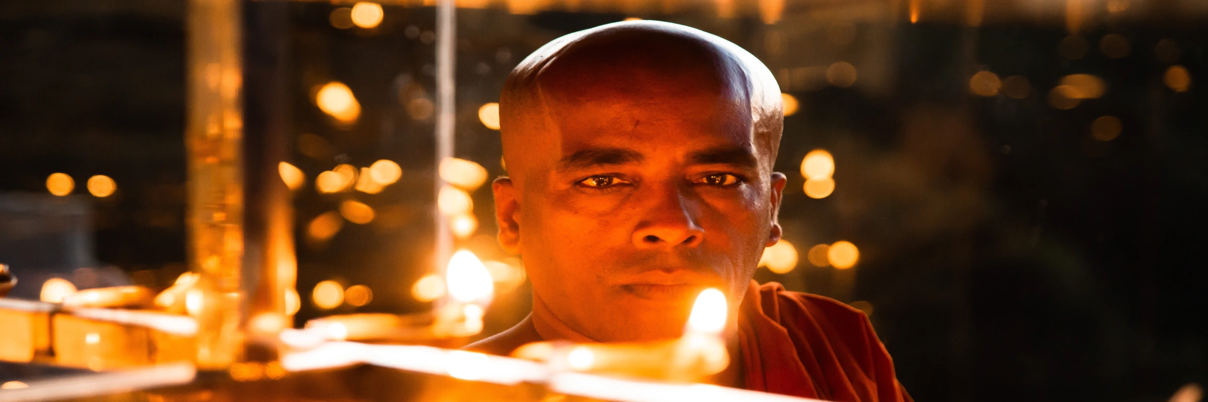 Ein buddhistischer Mönch blickt während einer traditionellen Pooja-Zeremonie in Sri Lanka hinter leuchtenden Öllampen hervor.