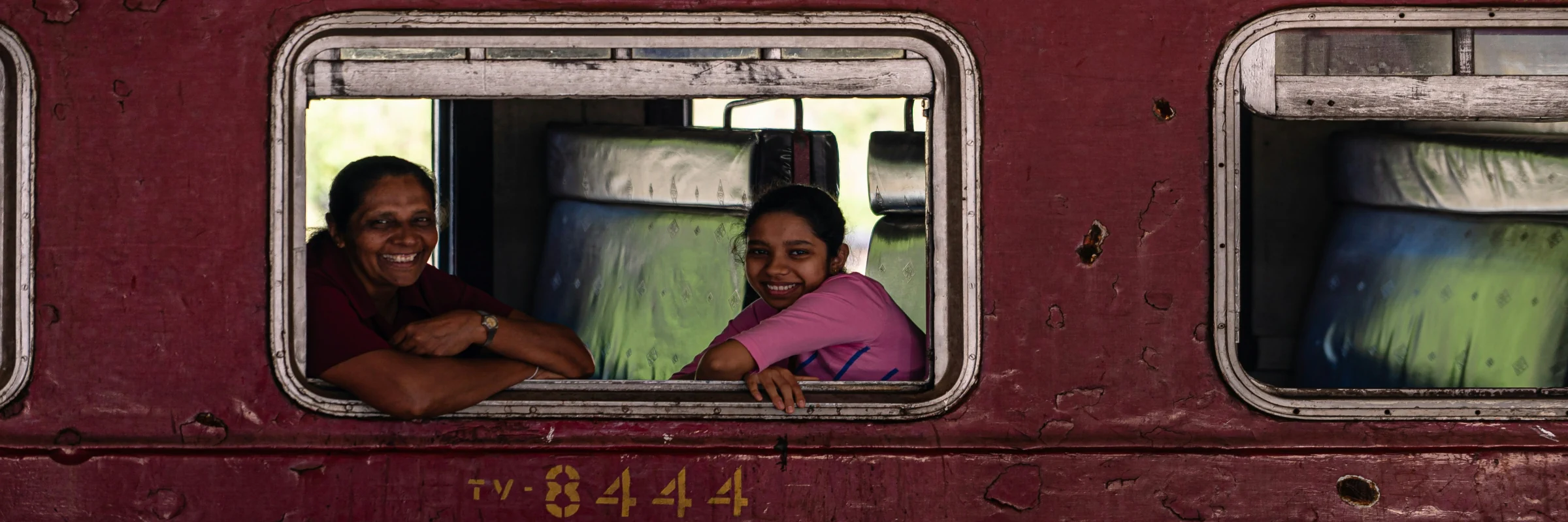 Zwei lächelnde einheimische Frauen blicken aus dem Fenster eines roten Zuges während der Zugfahrt im Hochland von Sri Lanka.